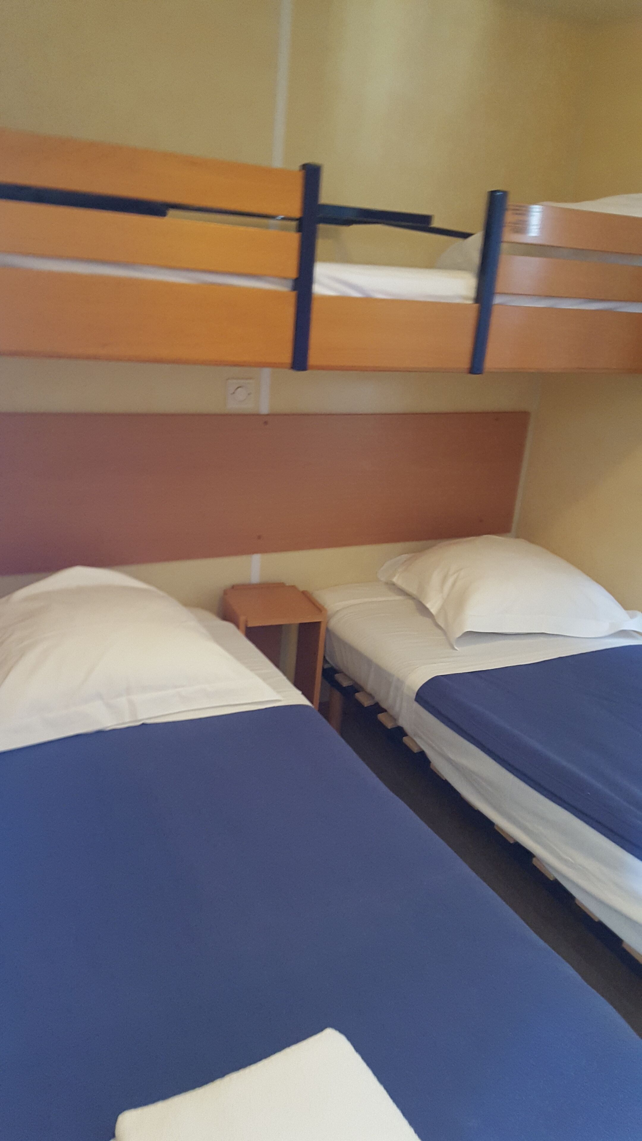 A dormitory room with two beds, a small wooden nightstand, and a bunk bed above.