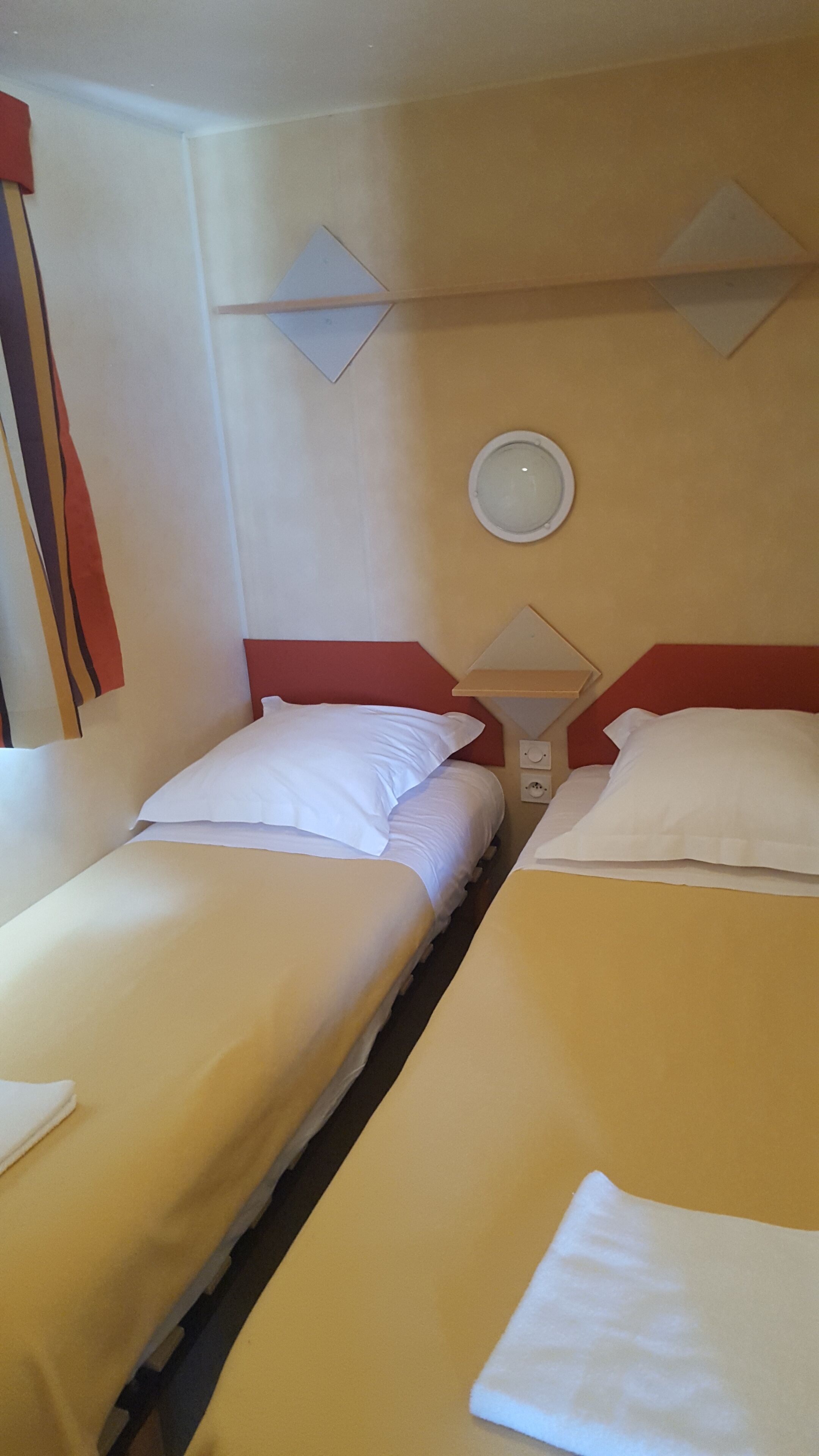 A hotel room with two single beds, each with white bedding and red headboards. There is a wall-mounted light fixture above the beds and a small shelf with a mirror and a clock.