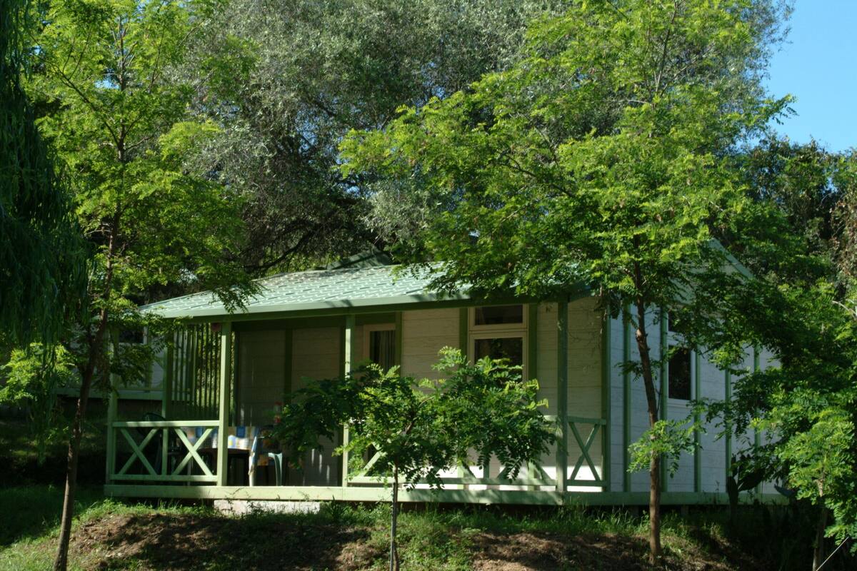 A small, single-story building with a green roof and white walls, surrounded by trees.