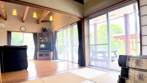 A spacious room with tatami mats, a large window with a view of the outdoors, a wooden ceiling, and a television. - Cottage Muinochichi (Motobu)