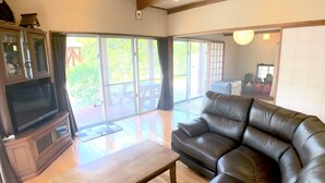 A spacious living room with a leather sofa, a television on a wooden cabinet, and large sliding glass doors leading to an outdoor patio. - Cottage Muinochichi (Motobu)