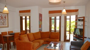A living room with a sectional sofa, a coffee table, a TV stand, and a dining area with chairs and a table. - Apartment DC01 - Kalypso (Kouklia)
