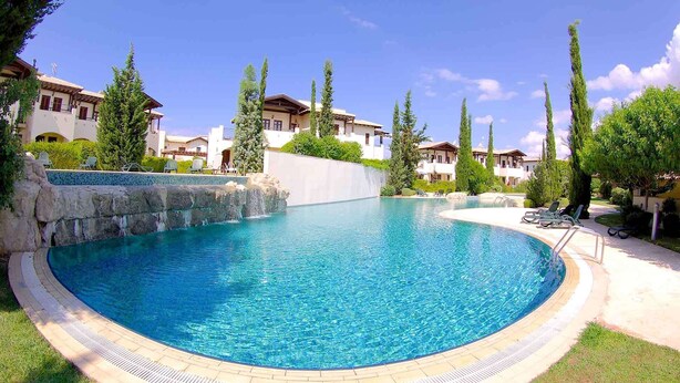 A large, curved swimming pool with a stone waterfall feature, surrounded by lush greenery and modern villas. - Apartment DC01 - Kalypso (Kouklia)