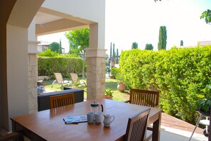 Una mesa de madera con sillas, una jarra y vasos, ubicada en un espacio al aire libre cubierto con vista a un jardín y un seto.