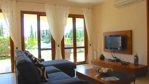 Ein Wohnzimmer mit einer Couch, einem Couchtisch, einem an der Wand montierten Fernseher und Blick ins Freie durch große Fenster.
