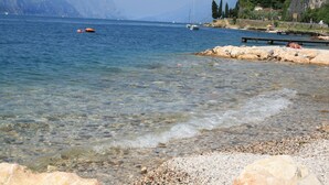 A clear lake with boats, pebbly shore, and mountains in the background. - Albergo Del Garda (Torri del Benaco)