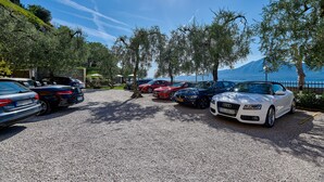 A parking area with multiple cars, including Audi and BMW models, near a body of water and a mountain in the background. - Albergo Del Garda (Torri del Benaco)