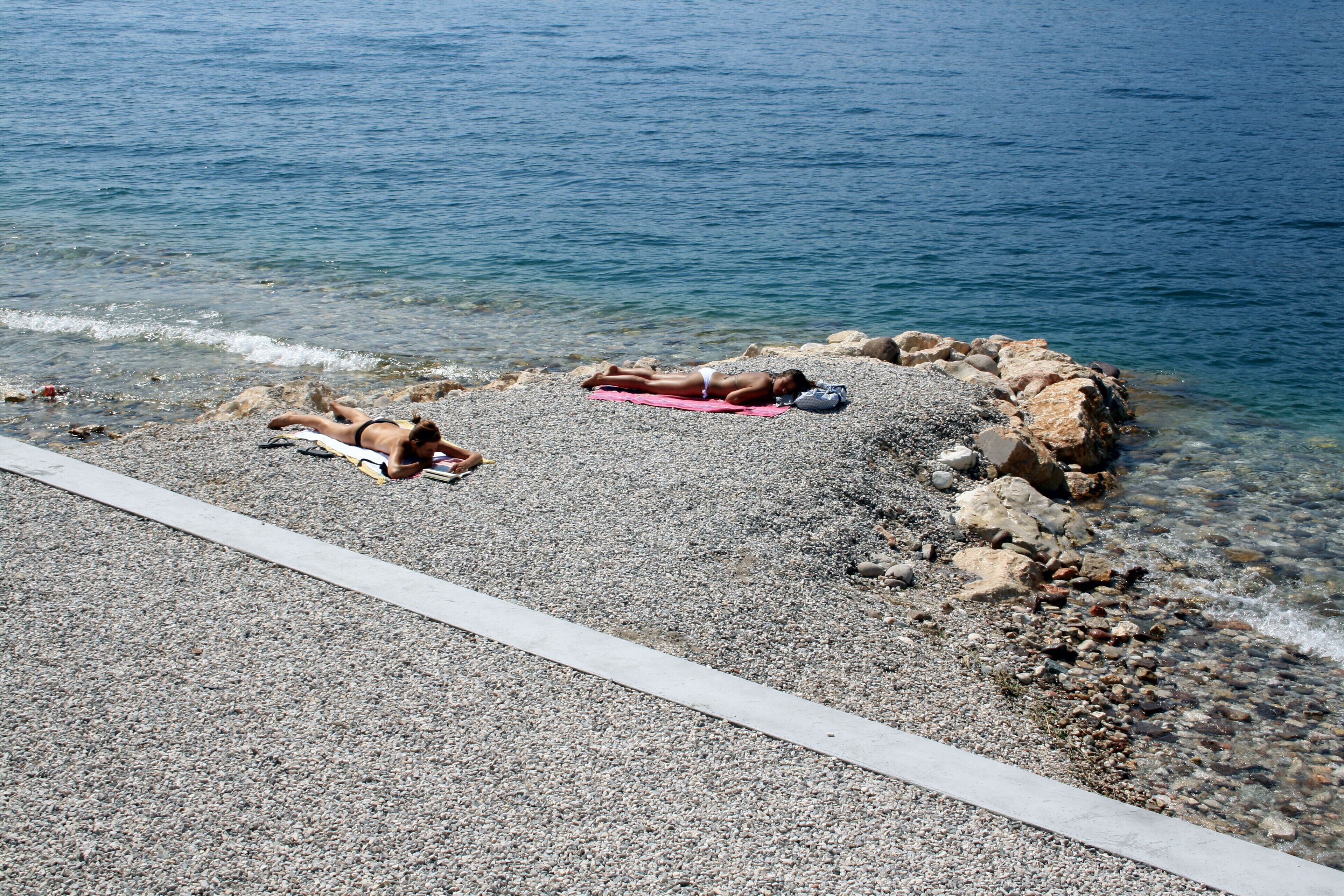 A pebbled beach with a person lying on a towel, a red towel, and a bag on the rocks.