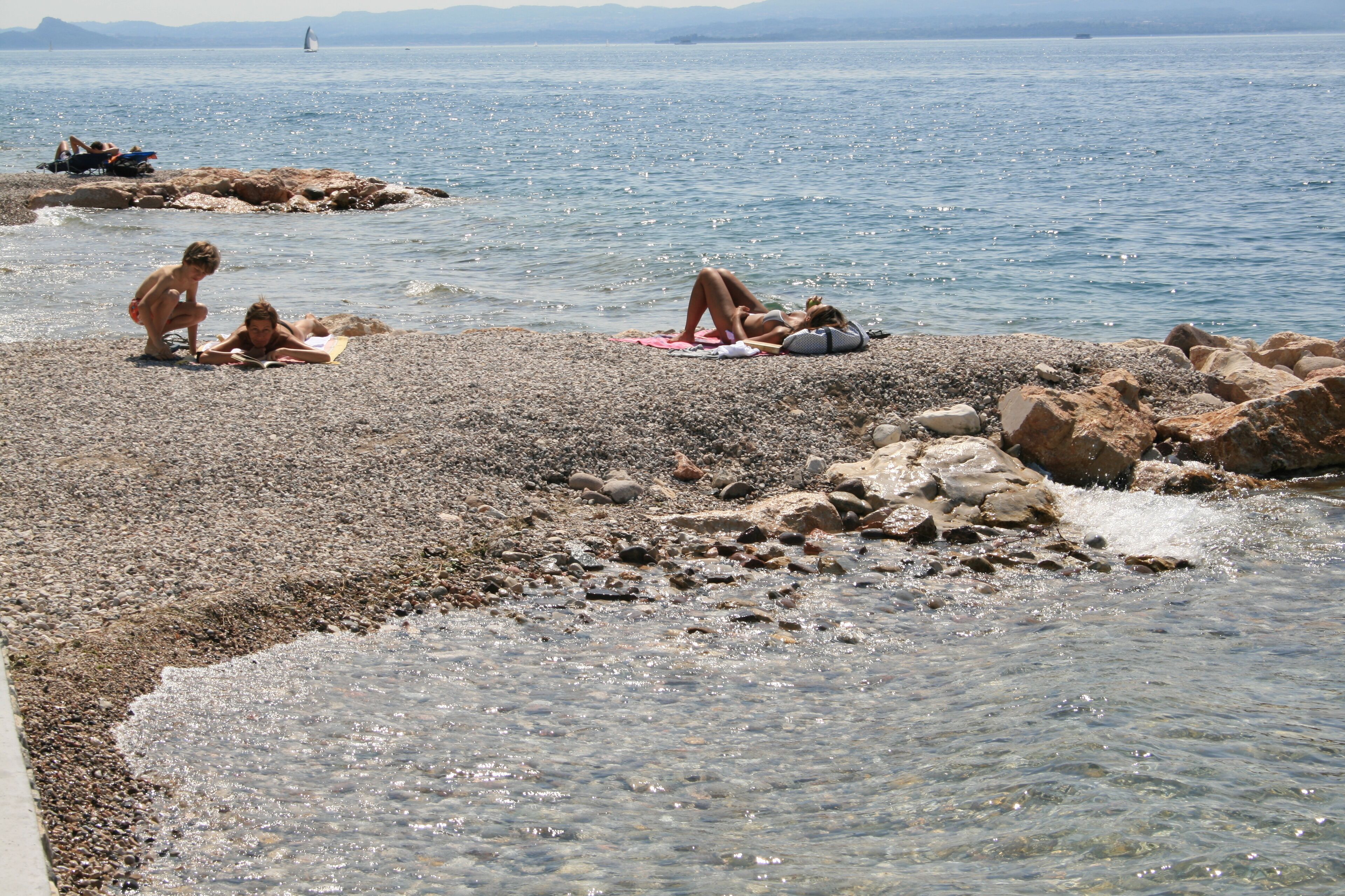 A pebbled beach with clear water, rocky outcrops, and people sunbathing.