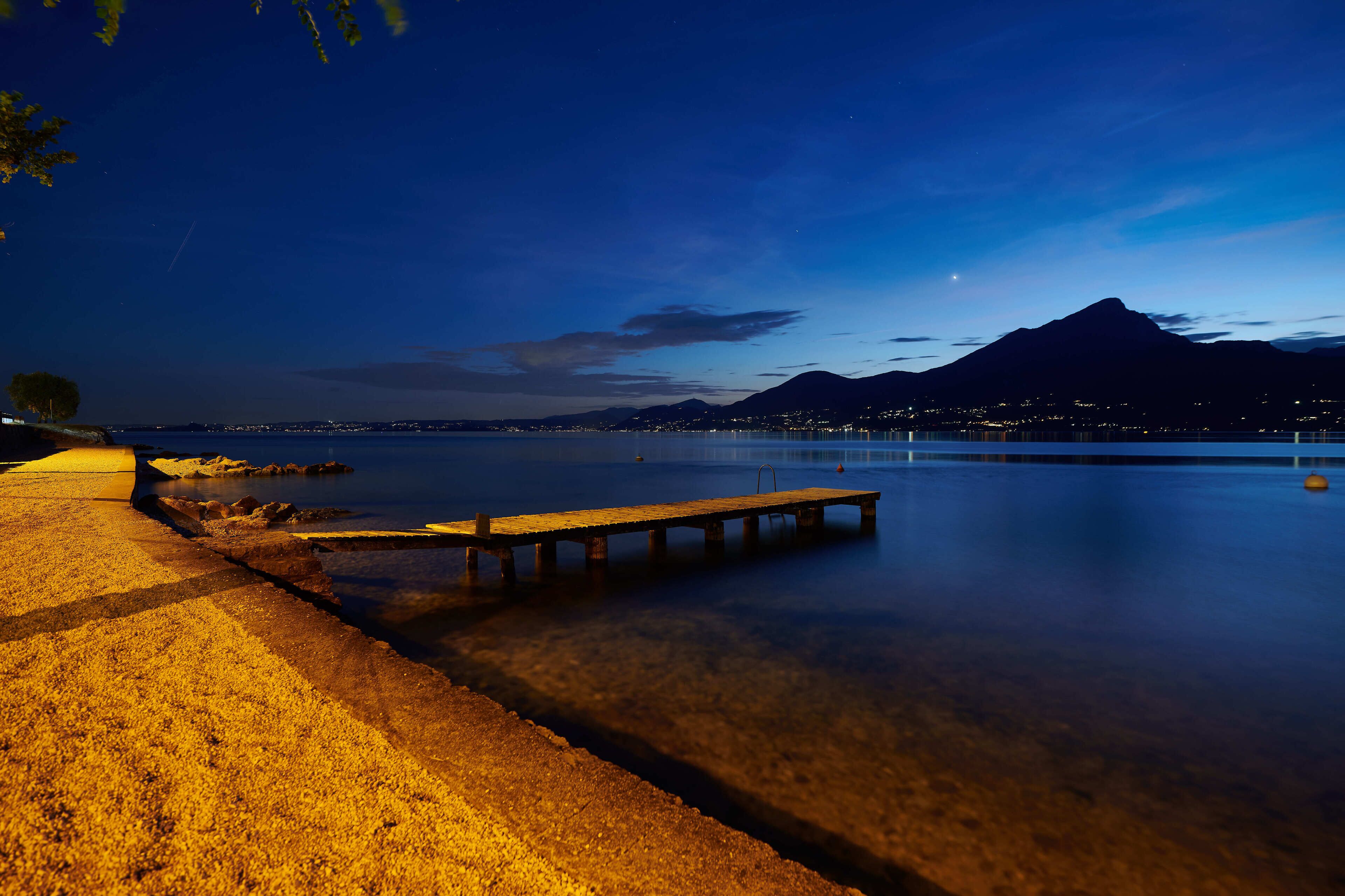 Vista nocturna de un lago tranquilo con un muelle de madera que se adentra en el agua, rodeado de una playa de arena y una montaña al fondo.