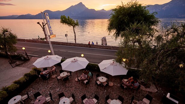 An outdoor seating area with tables and chairs, overlooking a body of water and mountains. - Albergo Del Garda (Torri del Benaco)