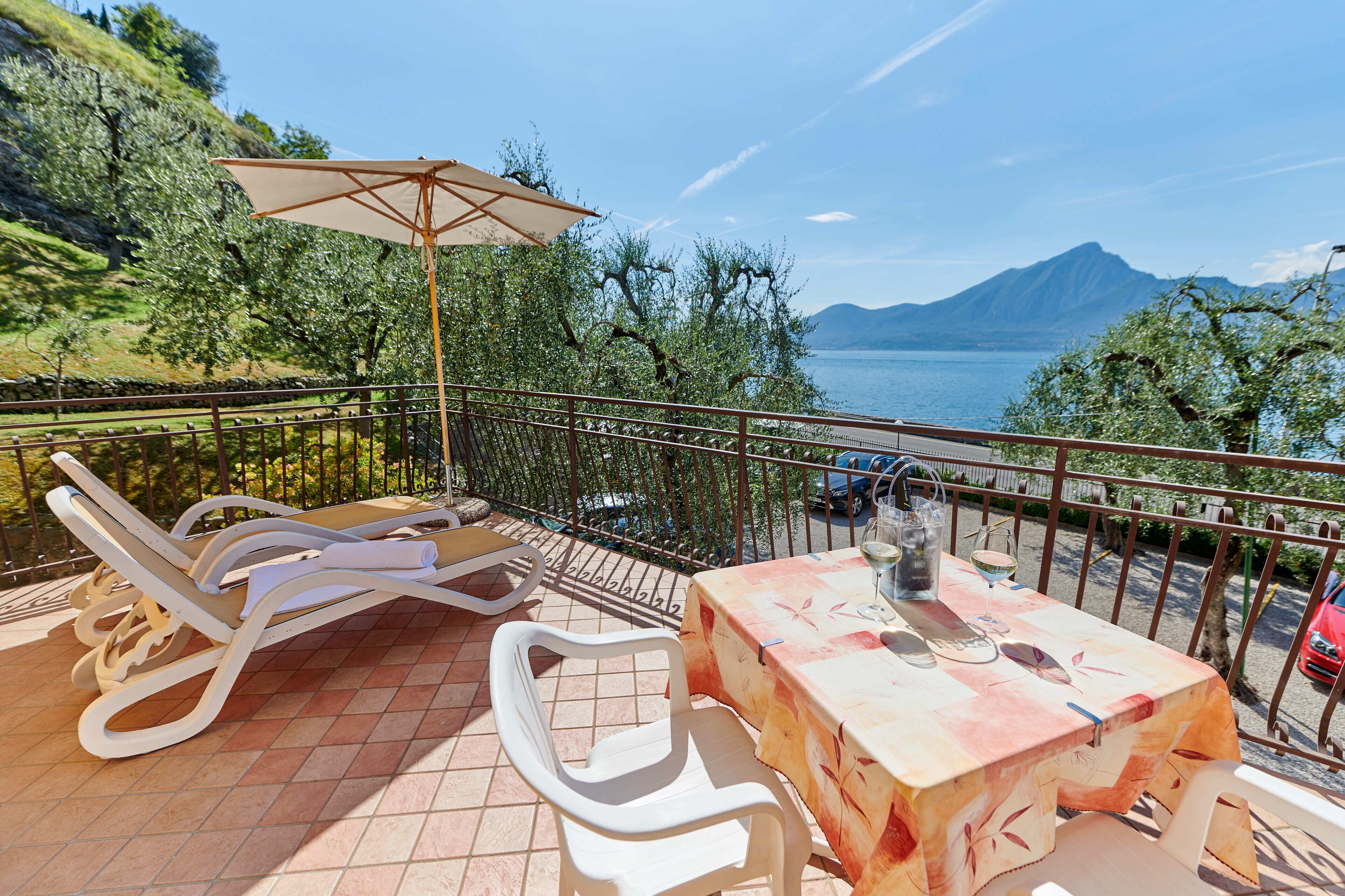 A balcony with a table and chairs, a table umbrella, and a view of a lake and mountains.