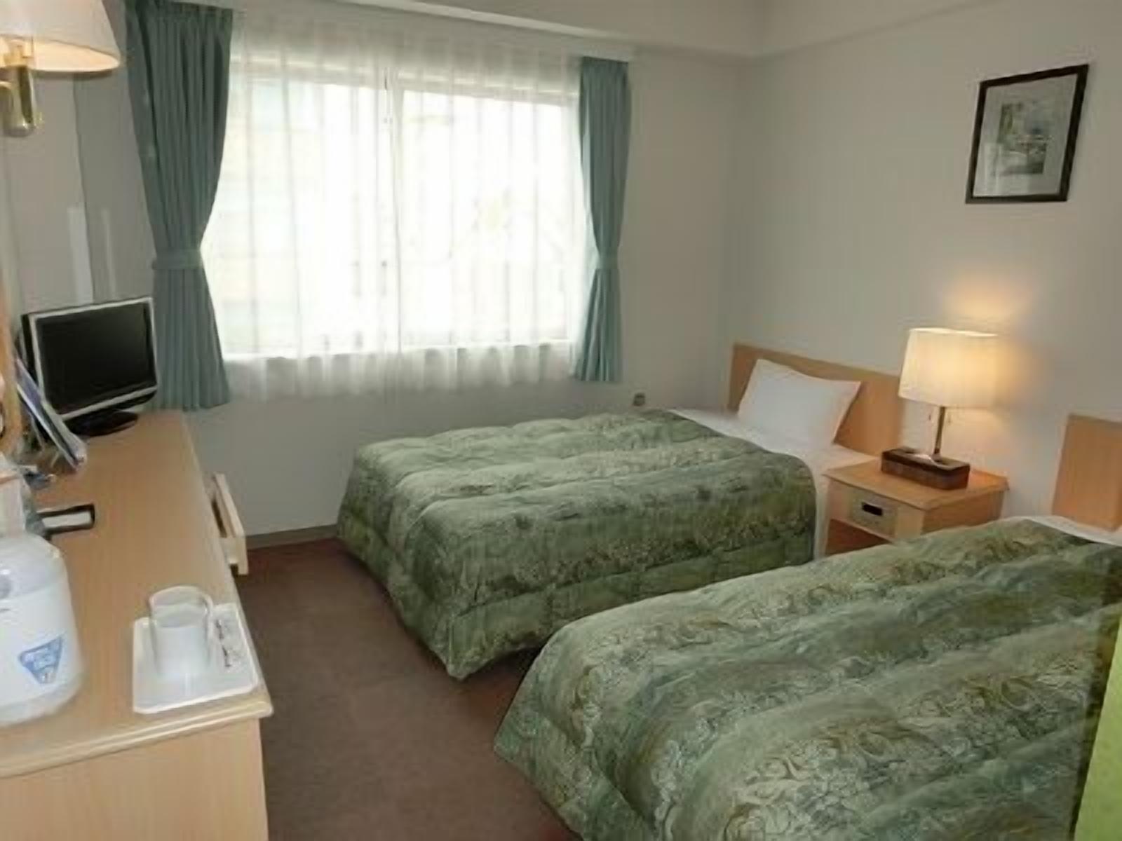 Habitació d'hotel amb dos llits, un escriptori amb un ordinador, una cafetera i una finestra amb cortines.