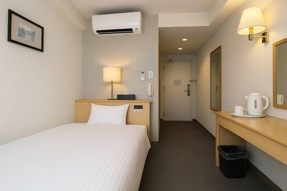A hotel room with a bed, bedside lamp, air conditioning unit, a mirror, a small table with a cup and a kettle, and a trash bin.