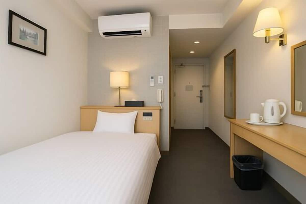 A hotel room with a bed, bedside lamp, air conditioning unit, a mirror, a small table with a cup and a kettle, and a trash bin.