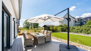 Een terras buiten met rieten meubilair en een grote parasol, met uitzicht op een verzorgd gazon en omliggende huizen.