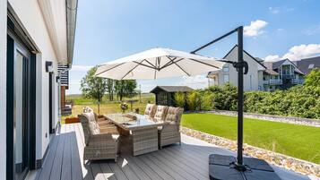 Eine Terrasse im Freien mit Korbmöbeln und einem großen Sonnenschirm, mit Blick auf einen gepflegten Rasen und angrenzende Häuser.