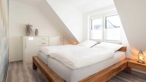 Een slaapkamer met een schuine kap, een bed met witte beddengoed, een nachtkastje met een lantaarn, en een raam met uitzicht op de omgeving.