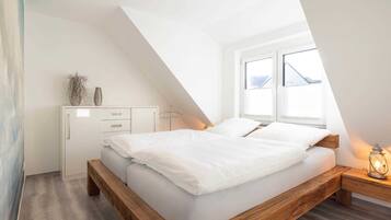 Ein Schlafzimmer mit schrägen Wänden, einem Bett mit weißer Bettwäsche, einem Nachttisch mit einer Laterne und einem Fenster mit Blick nach draußen.