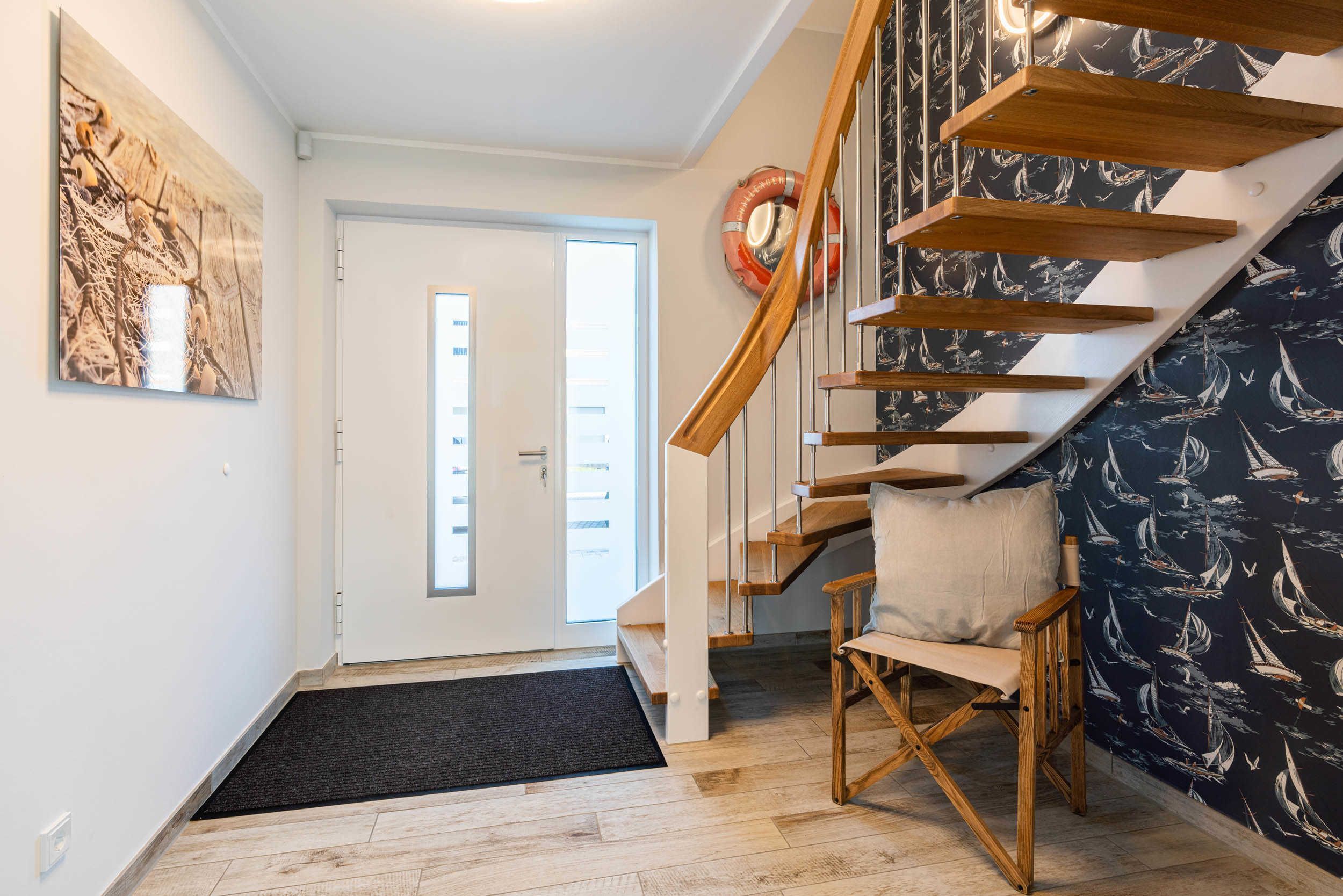 Una escalera de madera con escalones, una silla, una puerta con felpudo y una pared con papel tapiz de temática náutica.