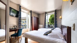 A hotel room with two beds, a desk with a chair, a TV mounted on the wall, and large windows with patterned curtains. - Hotel Beau Rivage (Aix-les-Bains)