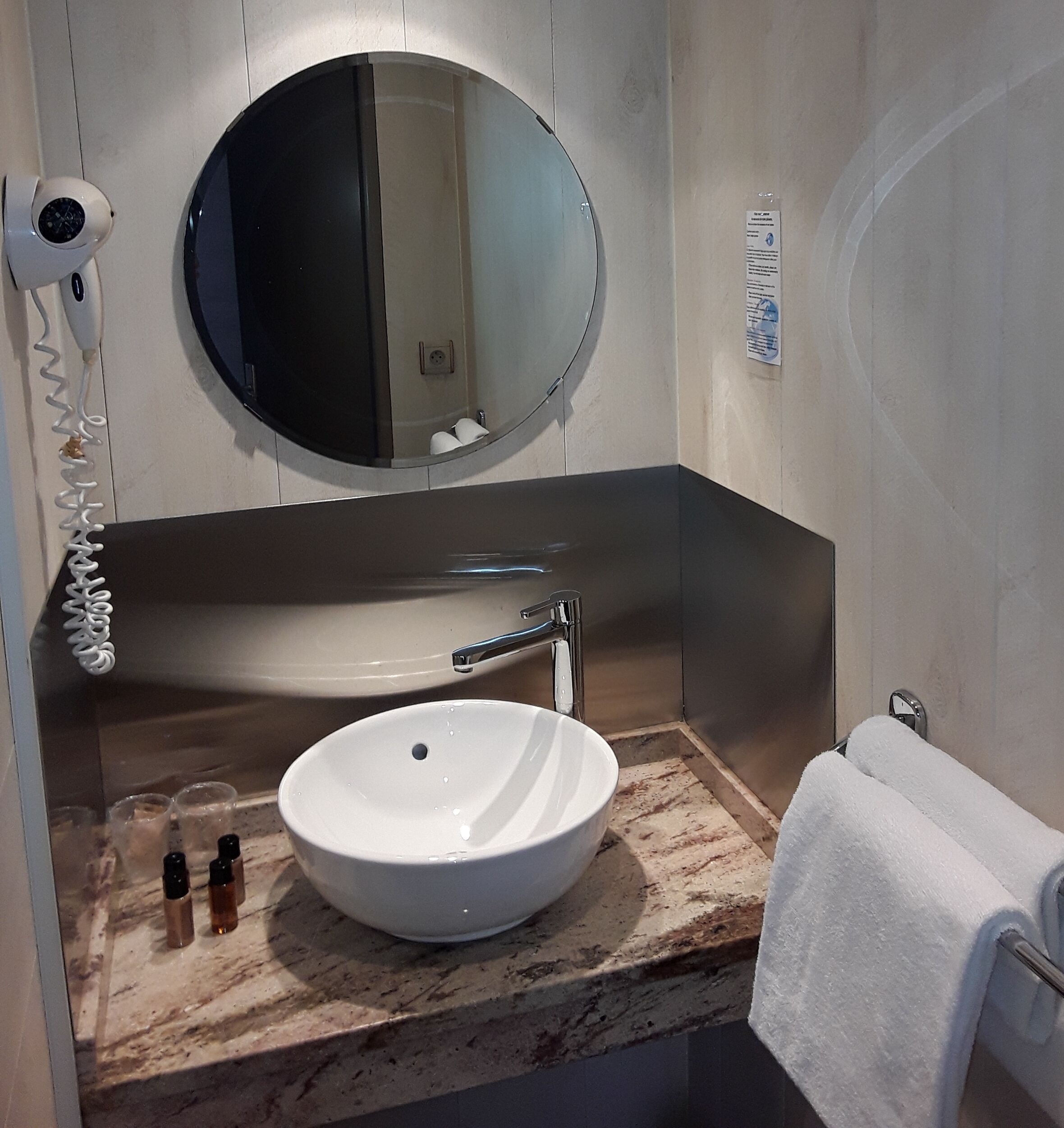 A bathroom with a round mirror, a white sink, a towel rack, and a hairdryer on the wall.