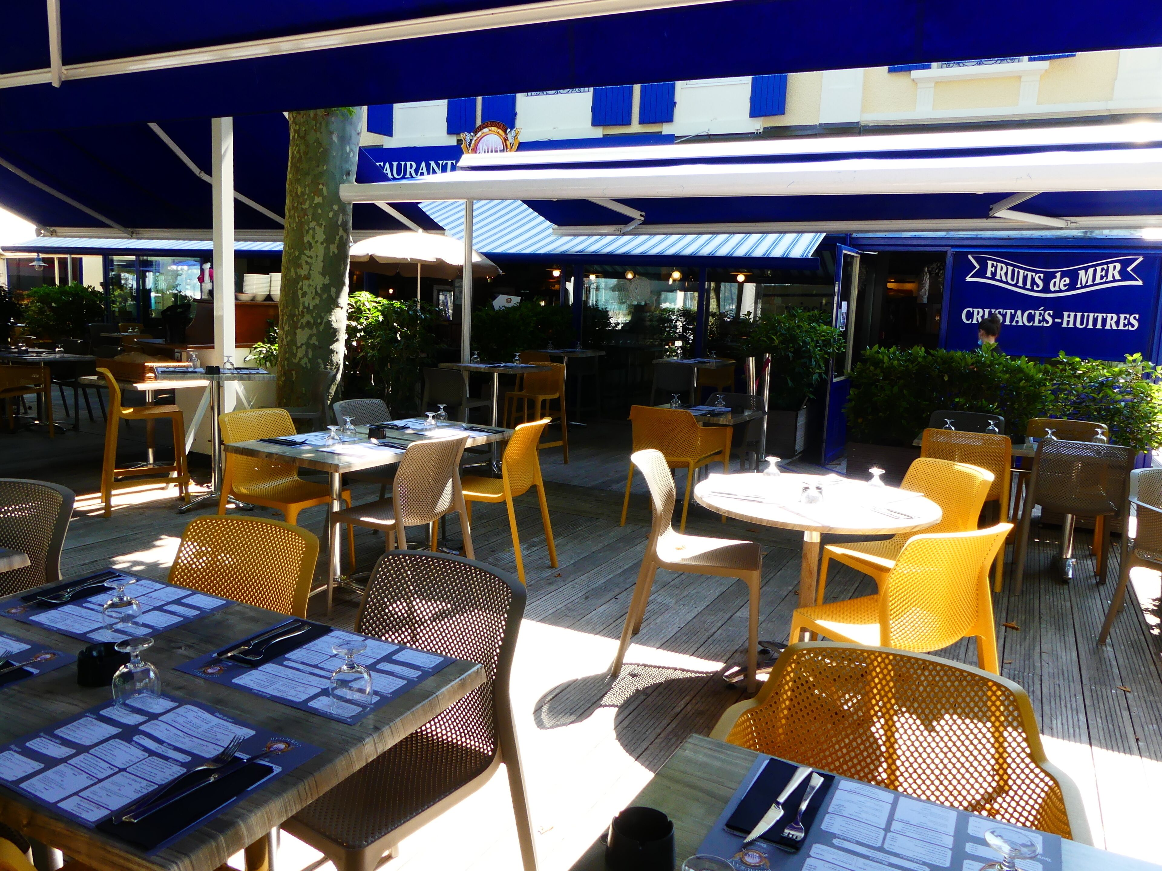 Un espace extérieur aménagé pour les repas, avec des tables et des chaises, un coin salon abrité et un panneau indiquant « FRUITS DE MER ».