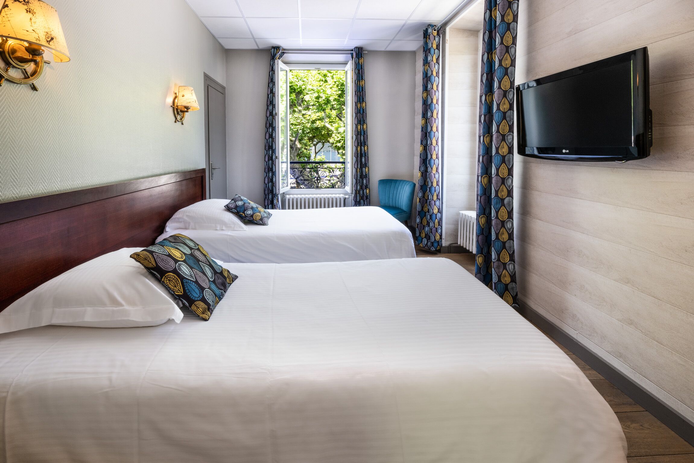 Ein Hotelzimmer mit zwei Betten, einem an der Wand montierten Fernseher und einem Fenster mit Vorhängen.