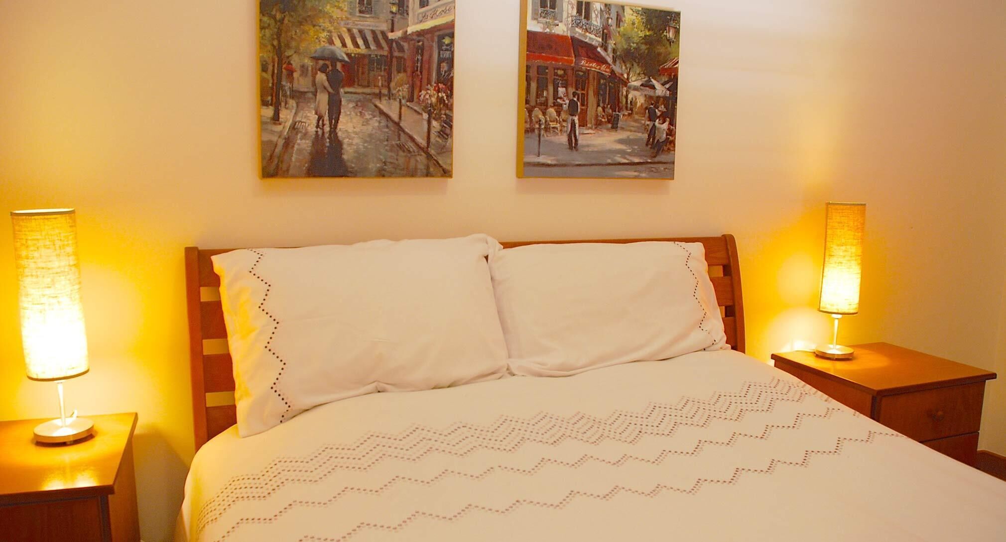 A hotel room with a bed, two paintings on the wall, and two bedside lamps.