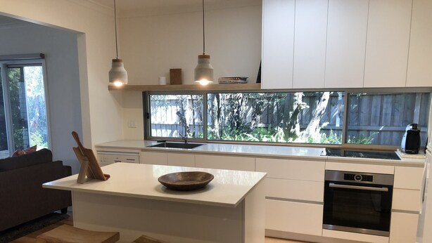 A modern kitchen with a large island, wooden stools, a built-in oven, and a view of the backyard through a large window. - Torquay - SpringHaven Beach House (Torquay)