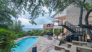 Une piscine entourée de chaises longues, une terrasse couverte et un escalier en pierre menant à un balcon du deuxième étage, d’où l’on a vue sur une étendue d’eau.