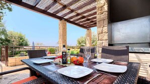An outdoor dining area with wicker tables, stone pillars, and a view of the landscape. - Villa HG01 - Madelini (Kouklia)