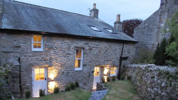 A stone house with two windows illuminated from the inside, a gravel path leading to the entrance, and a stone wall to the right.