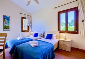 A room with two beds, a chair, a nightstand, a window, and a painting on the wall. - Villa 98 - Helidoni (Kouklia)