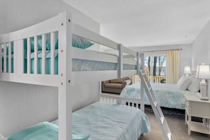Habitación de hotel con literas, un sofá, un escritorio con lámpara y vistas a árboles.