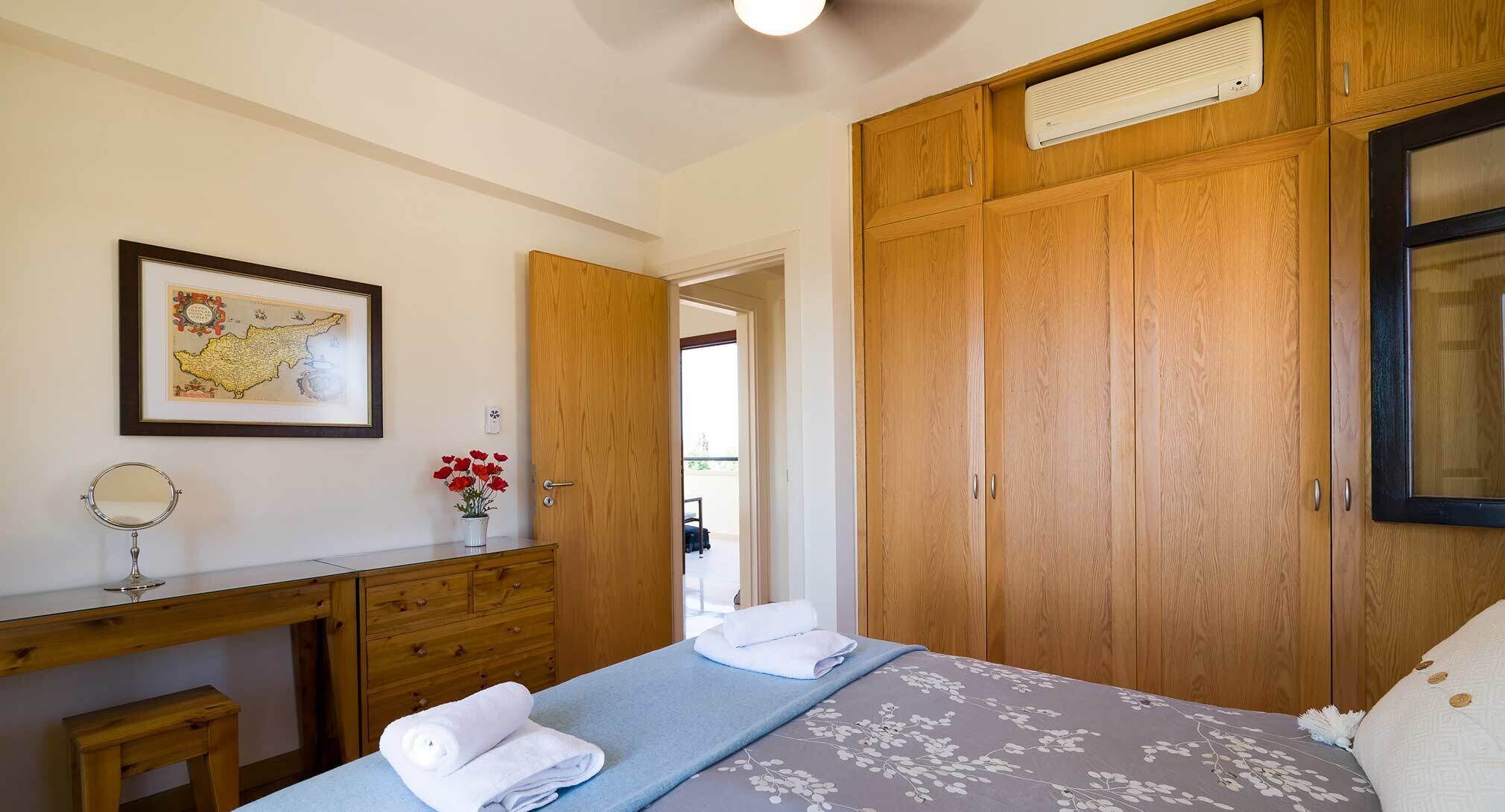 Un dormitorio con cama, armario de madera, escritorio con espejo, mapa enmarcado y un florero con flores rojas.