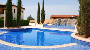 Una piscina con mosaicos azules, rodeada de follaje y un edificio con tejado de baldosas.