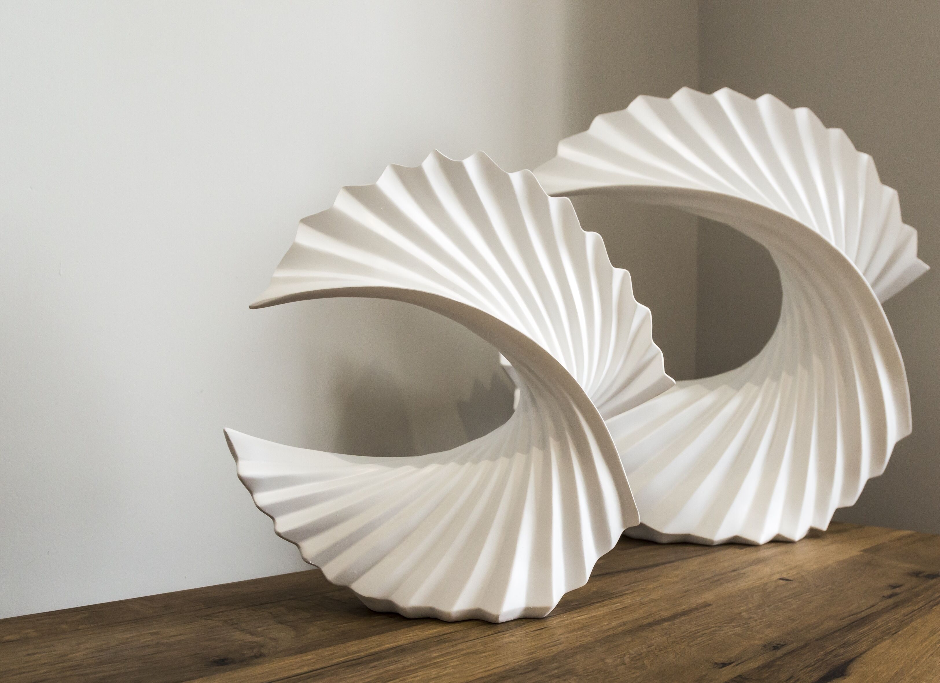 Una escultura abstracta blanca, con forma de volutas, sobre una superficie de madera frente a una pared lisa.