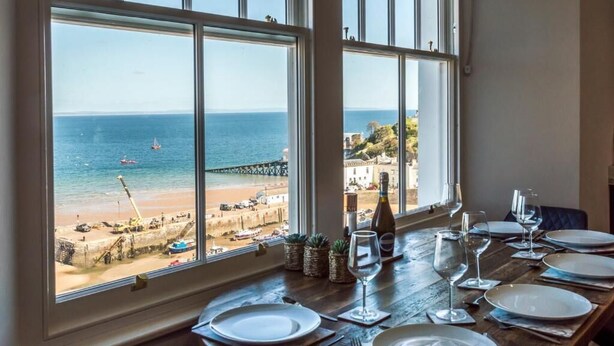 A dining table set for four with a view of the beach and pier through large windows. - Lady Tenby - 2 Bedroom Luxury Apartment (Tenby)