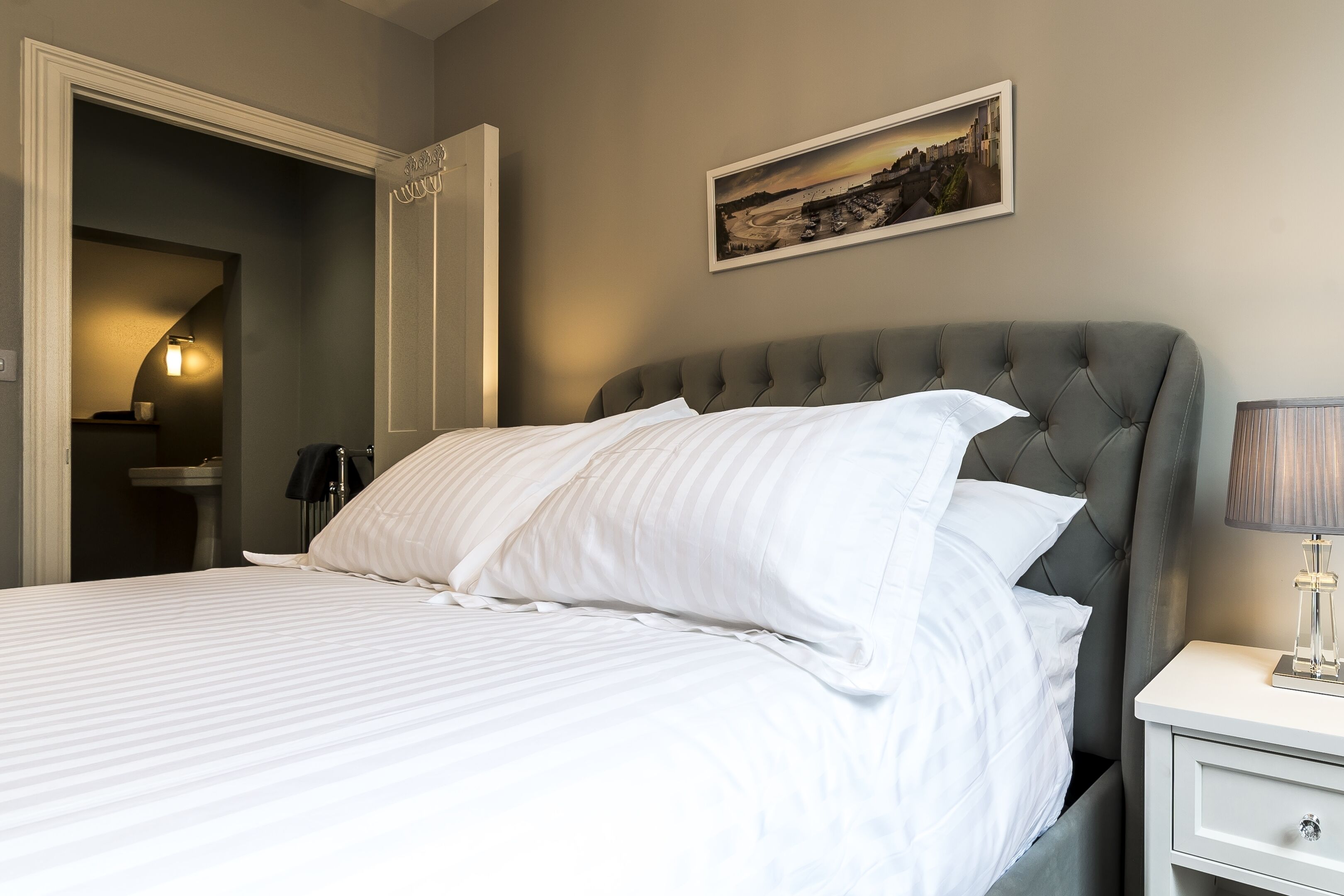 A neatly made bed with white linens, a grey upholstered headboard, and a bedside table with a lamp.