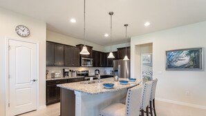 A modern kitchen with dark cabinets, a stainless steel refrigerator, a microwave, and a large island with blue bowls. There are pendant lights hanging from the ceiling and a clock on the wall. - Fascinating 5 BR Home Near Disney 8931 (Kissimmee)