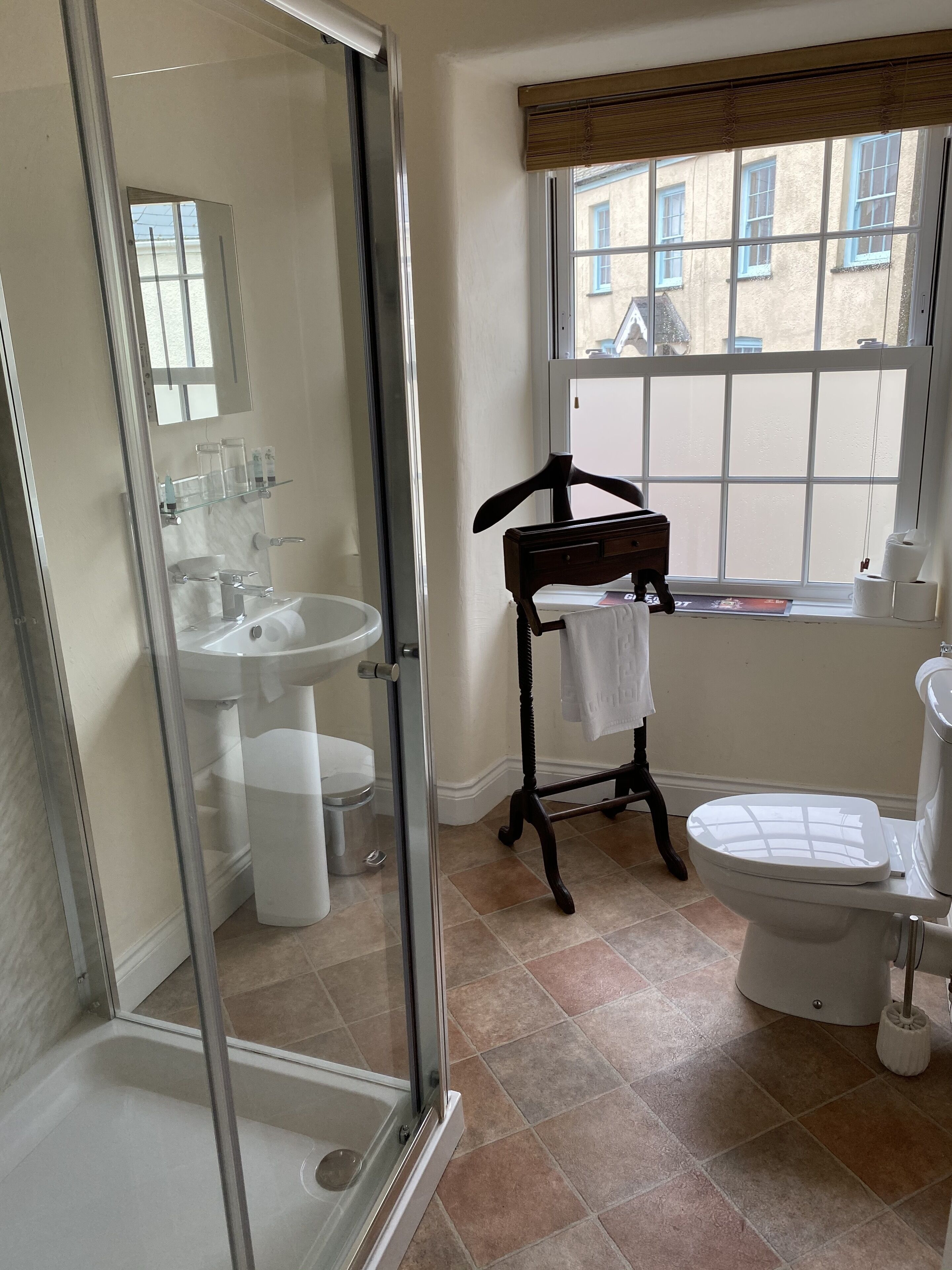 Ein Badezimmer mit Duschkabine aus Glas, Waschbecken, Toilette und Fenster mit Blick auf Gebäude.