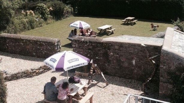 A patio area with picnic tables and an umbrella, overlooking a green landscape with hills. - The Poltimore Inn (South Molton)