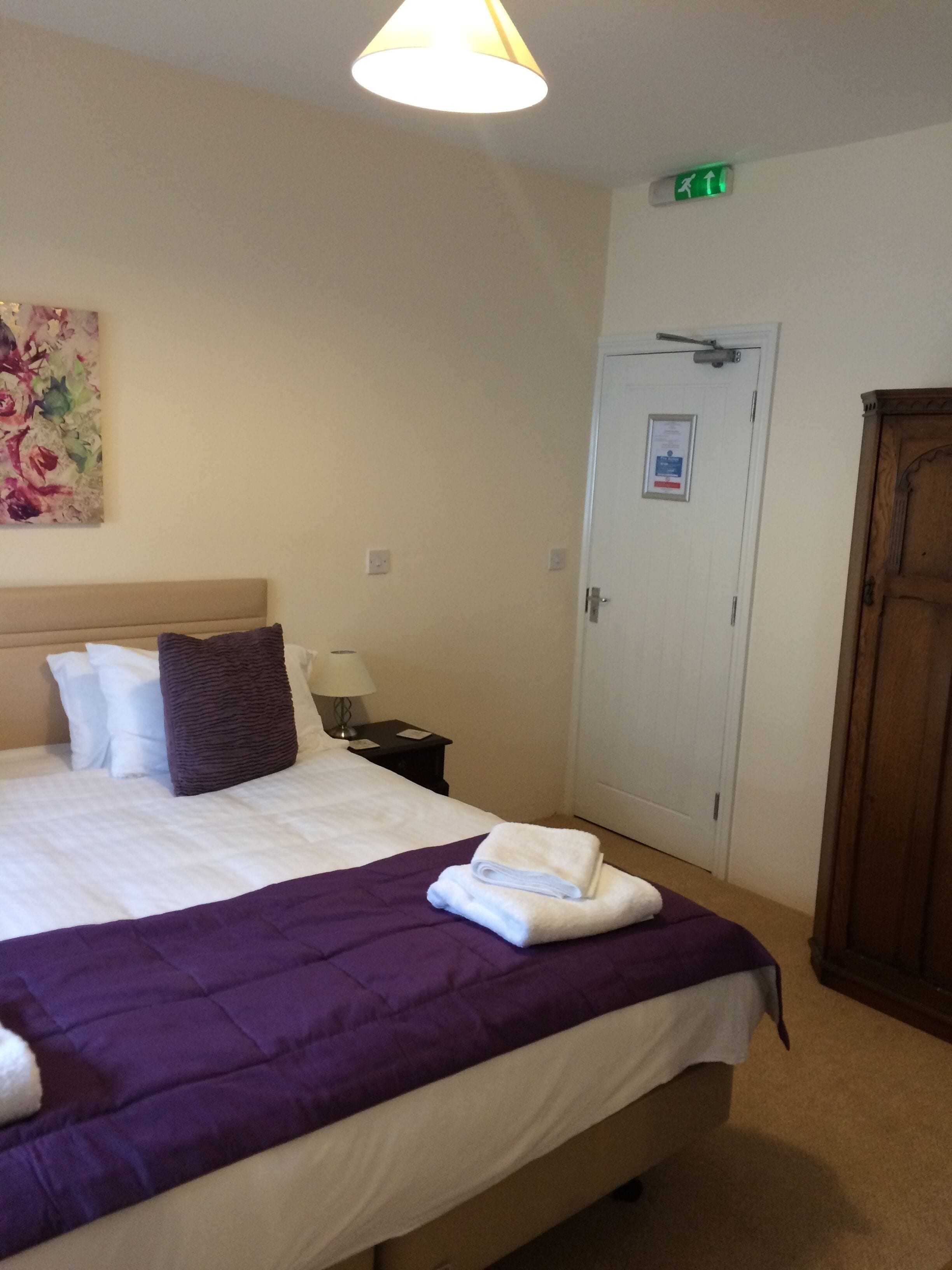 Una habitación de hotel con cama, mesita de noche, lámpara, un cuadro, una puerta y señal de salida de emergencia.