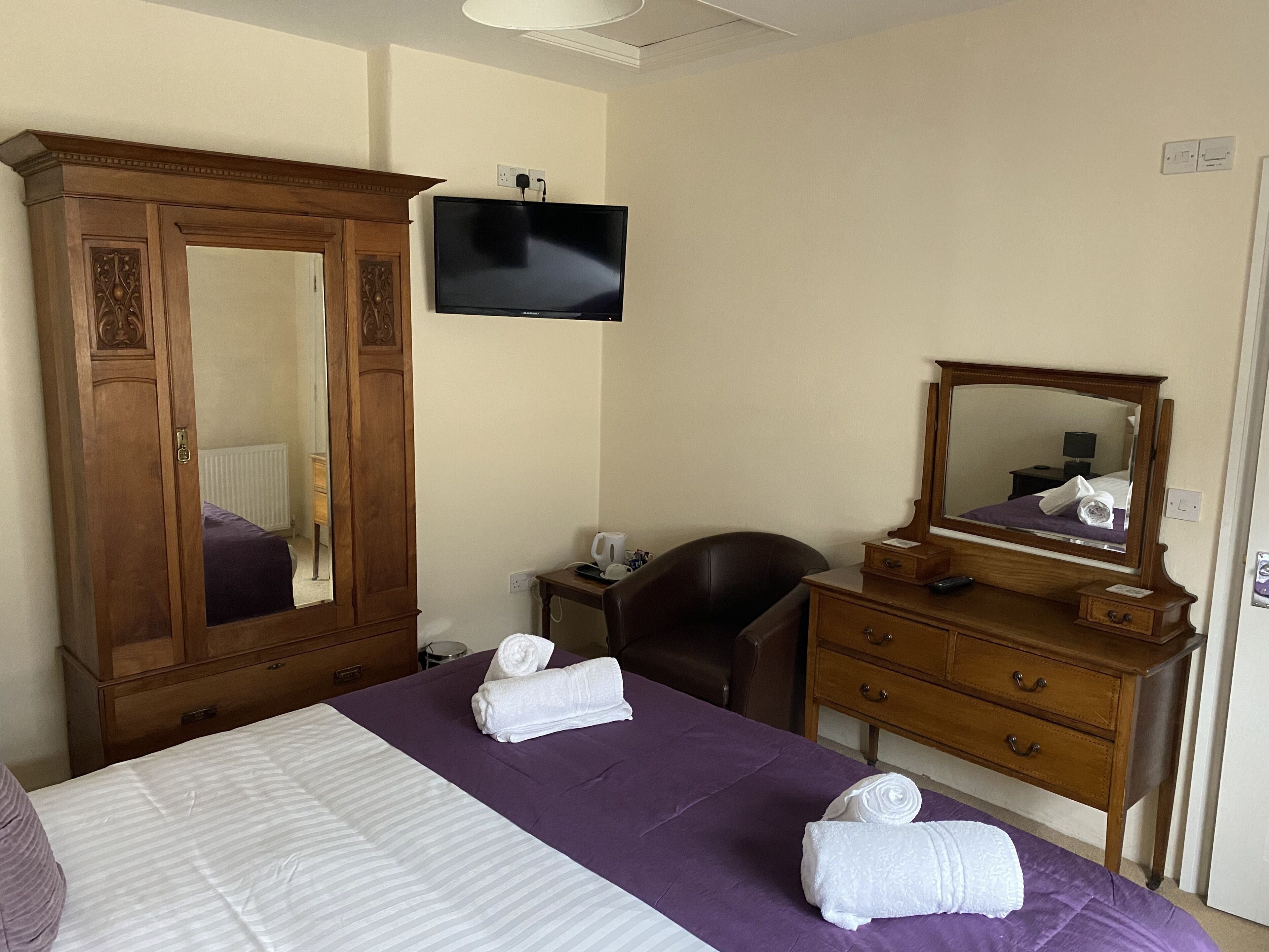 A hotel room with a large bed, a wooden wardrobe, a desk with a chair, a flat-screen TV mounted on the wall, and a mirror above the dresser.