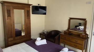 A hotel room with a large bed, a wooden wardrobe, a desk with a chair, a flat-screen TV mounted on the wall, and a mirror above the dresser.
