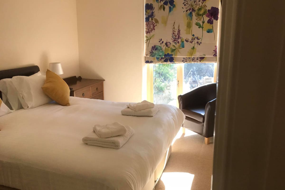 A neatly made bed with white linens and folded towels, a bedside lamp, and a floral curtain.