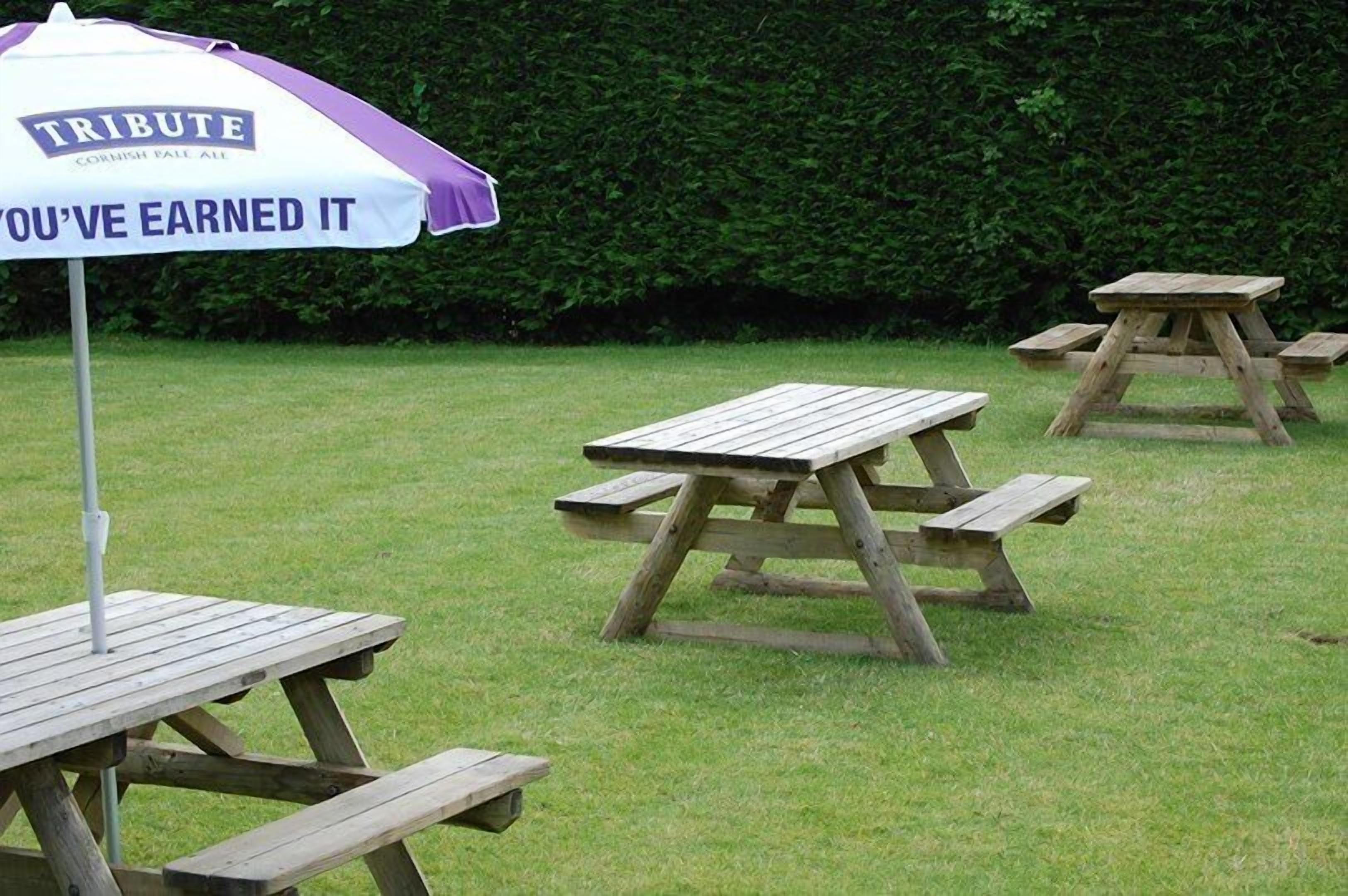 A picnic area with wooden tables and benches, an umbrella, and a hedge in the background.
