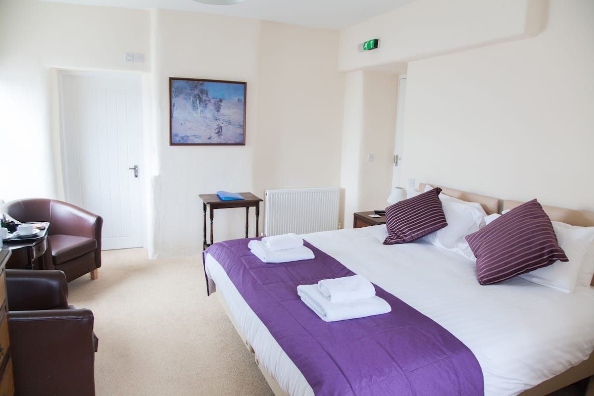 A hotel room with a bed, a desk, a chair, a radiator, and a painting on the wall.