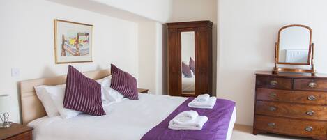A neatly made bed with a purple bedspread and white linens, a wooden dresser with a mirror, and a framed picture on the wall.